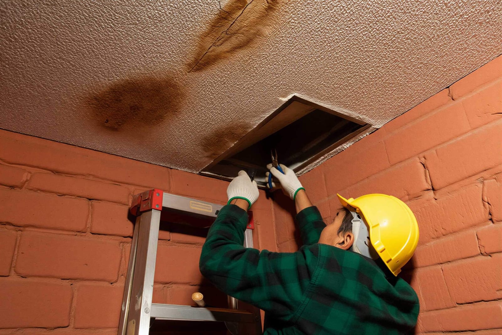 A technician inspecting a roof leak at a water-stained ceiling on a ladder
